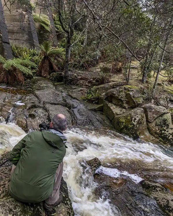 A photographer wearin a dark green rain jacket is squatting on a rock beside the Cascade River just below the Mt Paris dam wall. He is using a camera with a large lens on a tripod.